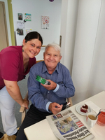 Ein älterer Mann sitzt an einem Tisch mit einer Zeitung und einem Stück Kuchen. Neben ihm steht eine lachende Frau in medizinischer Bekleidung. Beide scheinen eine angenehme Zeit zusammen zu verbringen. Auf dem Tisch steht auch eine Tasse Kaffee.
