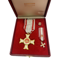 Eine Medaille in einer roten Präsentationsbox, die eine goldene Kreuzform mit einem roten und weißen Band zeigt. Neben der Medaille sind ein weiteres kleines Kreuz und ein Ansteckbutton sichtbar. Der Innenraum der Box ist mit rotem Stoff ausgekleidet.