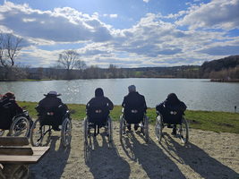 Personen im Rollstuhl sitzen am Ufer eines ruhigen Sees unter einem blauen Himmel.