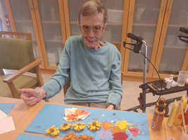 Eine ältere Person sitzt an einem Tisch und arbeitet an einem Kunstprojekt mit gefärbten Papierblumen. Vor ihr sind bunte Blumen auf einem Blautonkarton angeordnet. Der Raum wirkt hell und einladend, mit weiteren Malutensilien auf dem Tisch.