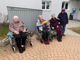 Vier ältere Menschen in Rollstühlen sitzen draußen auf einem gepflasterten Weg.