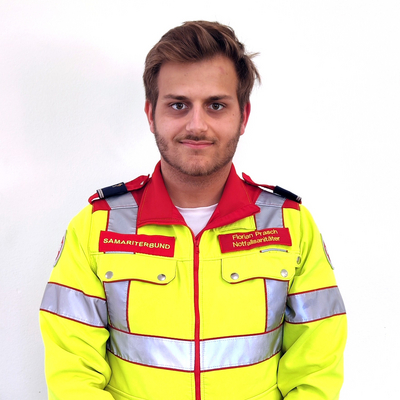 Ein junger Mann in einer auffälligen, gelben Einsatzjacke mit reflektierenden Streifen steht vor einem weißen Hintergrund. Die Jacke ist mit dem Schriftzug 