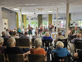 Eine Gruppe von Musikern spielt auf der Bühne in einem Gemeinschaftsraum. Vor ihnen sitzt ein Publikum älterer Menschen, einige in Rollstühlen. Die Atmosphäre ist fröhlich und die Musik unterhält die Anwesenden.