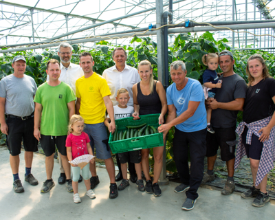 Gruppenfoto in Gewächshaus bei Simmeringer Bauern, mehrere Erwachsene, aber auch Kinder halten grüne Kiste mit Gurken, im Hintergrund Pflanzen