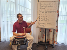 Ein Mann spielt Schlagzeug und erklärt Musiknotationen an einem Flipchart.