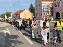 Eine lange Gruppe von Menschen, darunter ältere Personen und Kinder, geht auf einer Straße. Einige nutzen Rollatoren oder Gehhilfen. Eine Person trägt eine Leuchtweste. Im Hintergrund sind Häuser und ein farbenfrohes Wandgemälde zu sehen.