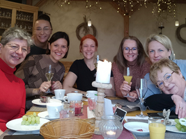 Eine Gruppe von sieben Frauen sitzt an einem festlich gedeckten Tisch mit verschiedenen Speisen und Getränken. Der Tisch ist mit einer Kerze und dekorativen Lichtern geschmückt. Alle Frauen lächeln und posieren fröhlich für das Foto.