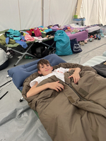 Ein Junge liegt in einem Schlafsack und lächelt. Kleidung ist im Hintergrund gestapelt.