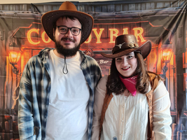 Zwei Personen in Cowboy-Kleidung stehen neben einem großen Topf Chili.