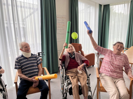 Senioren sitzen in Rollstühlen und heben bunte Gegenstände zum Spielen.