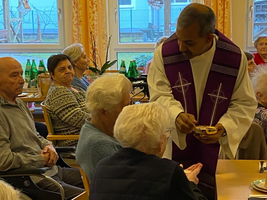 Ein Priester reicht älteren Menschen in einem Gemeinschaftsraum eine Schale.