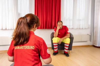 Zwei Menschen im Gespräch, einer in rotem Shirt, der andere in gelber Hose.