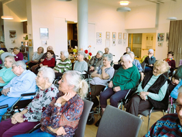 Eine Gruppe älterer Menschen sitzt in einem hellen Raum. Einige tragen Brillen und unterschiedliche Kleidung. Sie schauen aufmerksam nach vorne, in einer Atmosphäre der Gemeinschaft und Interaktion. Im Hintergrund sind Bilder an den Wänden zu sehen.