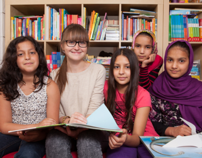 5 Kinder sitzen mit Buch auf Couch und lächeln in Kamera, im Hintergrund Bücherregal mit vielen Büchern