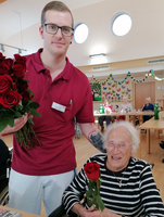 Ein junger Mann überreicht einer älteren Dame eine rote Rose in einem Gemeinschaftsraum.