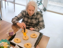 Eine ältere Person sitzt an einem Tisch und bestreicht Teiglinge auf einem Backblech mit einem Pinsel. Vor ihr steht eine Tasse und in der Nähe ist eine Schüssel mit Zutaten sichtbar. Hintergrund mit natürlichem Licht durch Fenster.