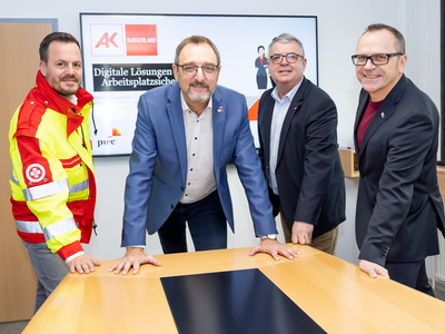 Vier Männer stehen um einen Tisch und diskutieren digitale Lösungen für Arbeitsplätze.
