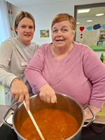Zwei Frauen stehen über einem großen Topf mit einer Suppe. Eine Frau rührt mit einem langen Löffel in der Suppe, während die andere lächelt. Beide wirken freundlich und freuen sich am gemeinsamen Kochen in einer hellen Küche.