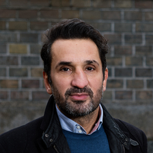 Ein Mann (Fadi Merza) steht draußen vor einer Ziegelmauer, 
