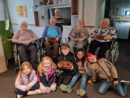 Ältere Menschen und Kinder sitzen zusammen und halten Obstschalen in einem Gemeinschaftsraum.
