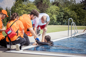 Ein Team von Rettungsschwimmern in orangefarbenen T-Shirts leistet Hilfe an einem Schwimmbecken. Eine Person im Wasser wird von zwei Rettungskräften an der Hand gezogen, während ein weiterer Helfer bereit steht. Im Hintergrund sind Bäume und eine Wiese zu sehen.