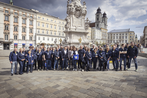 Gruppe von Menschen in blauen Shirts steht vor einem Denkmal auf einem Platz.