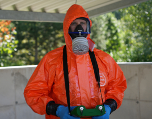 Person in orangen Schutzanzug und Maske, mit Strahlungsdetektor in der Hand