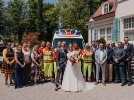 Hochzeitsfeier mit Brautpaar und Rettungskräften des ASBÖ im Freien; fröhliche Gesichter.
