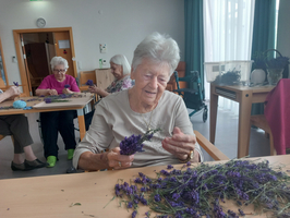 Eine ältere Frau sitzt an einem Tisch und bindet Lavendelsträuße. Im Hintergrund sind weitere Frauen, die ebenfalls mit Lavendel arbeiten. Die Szene spielt in einem hellen Raum mit großen Fenstern und einem freundlichen Ambiente.