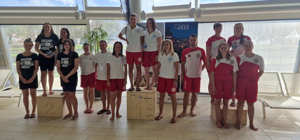 Gruppenfoto von Schwimmern in roten und weißen Outfits, mit Pokalen auf Podesten.