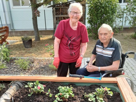 Ein älteres Paar steht neben einem Hochbeet mit frisch gepflanzten Blumen.
