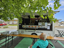 Ein Lebensmittelstand mit einem Verkäufer, der bestellte Speisen zubereitet. Vor dem Stand sitzt eine ältere Person in einem Rollstuhl. Auf einem Tisch liegen mehrere Teller bereit. Die Umgebung ist sonnig, unter einem Baum mit grünem Laub.