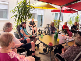 Eine Gruppe von älteren Menschen sitzt im Freien in einem Aufenthaltsbereich mit Sonnenschirmen. Einige verwenden Rollstühle, während andere in bequemen Stühlen sitzen. Pflanzen umgeben den Bereich. Ein Tisch ist mit Getränken und einem grünen Wasserbehälter gedeckt.