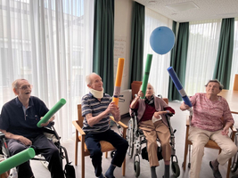 Ältere Menschen spielen mit Bällen und Schaumstoffröhren in einem hellen Raum.