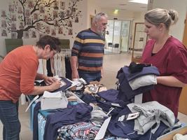 Drei Personen sortieren Kleidung an einem Tisch in einem Innenraum.
