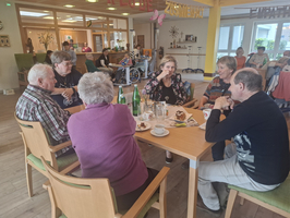 Eine Gruppe von sieben älteren Menschen sitzt an einem Tisch in einem Gemeinschaftsraum. Sie unterhalten sich und genießen Getränke und Kuchen. Im Hintergrund sind weitere Personen und Einrichtungen zu sehen, die eine freundliche, einladende Atmosphäre schaffen.
