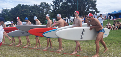 Sechs Sportler stehen mit Rettungsbrettern an der Startlinie eines Wettkampfs.