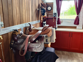 In einem Stallraum sind mehrere Sättel auf einem Tisch gestapelt. Die Wände sind aus Holz, und es gibt ein Fenster mit roten Vorhängen. Im Hintergrund steht eine Kücheneinheit mit roten Schränken. An der Wand hängt eine Uhr.