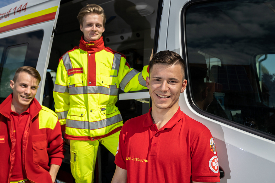 Drei Rettungskräfte lächeln vor einem Rettungswagen.