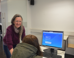 Foto aus dem Internetcafe, eine Frau von vorne steht neben Tisch und lächelt in Kamera, Klientin sitzt vor Computer Gesicht nicht erkennbar