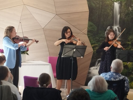 Drei Geigerinnen spielen Musik vor einem Publikum in einem hellen Raum.