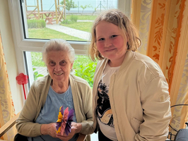 Eine ältere Frau sitzt an einem Tisch und hält eine bunte Origami-Blume in der Hand. Neben ihr steht ein jüngeres Mädchen, das lächelt. Im Hintergrund sind Fenster und ein Garten zu sehen.