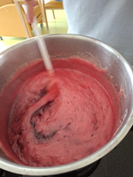 Ein runder Kochtopf mit einer schaumigen, pink-roten Masse wird mit einem Löffel verrührt. Im Hintergrund sind unscharfe Stühle und Tische zu sehen.