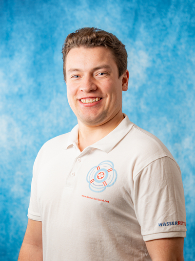 Junger Mann in einem hellen Polo-Shirt mit Wasserrettungslogo lächelt vor blauem Hintergrund.