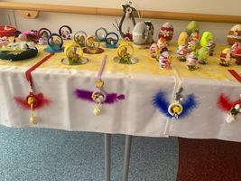 Auf einem Tisch sind verschiedene handgemachte Osterdekorationen ausgestellt. Darunter befinden sich bunte Eier, kleine Figuren und kreative Arrangements mit Federn und glitzernden Elementen. Die Farben sind fröhlich, typisch für die Osterzeit, und laden zur Feier ein.