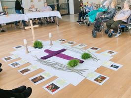 Ein Raum mit Senioren, die um ein zentrales, mit einer lila Decke und einem Kreuz dekoriertes Arrangement sitzen. Um das Arrangement liegen Bilder und Texte. Die Atmosphäre ist ruhig und nachdenklich.