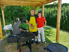 Zwei Personen stehen neben einem Grill unter einem überdachten Bereich im Garten.