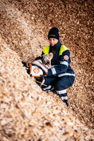 Eine Frau in Sicherheitskleidung kniet auf einem Holzschnitzel-Haufen und streichelt einen Hund, der ebenfalls mit einem Dienstgeschirr ausgestattet ist. Beide scheinen an einer Rettungsoperation beteiligt zu sein. Der Hintergrund besteht aus groben Holzschnitzeln.