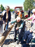 Eine Gruppe von Menschen, darunter ältere Personen, arbeitet gemeinsam im Freien an einem Holzstück. Eine Person im Rollstuhl bearbeitet das Holz mit einem Werkzeug, während andere assistieren. Im Hintergrund sind ein Traktor und mehrere Erwachsene zu sehen.