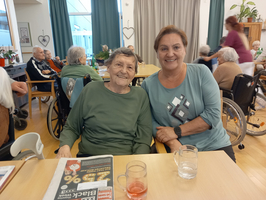 Eine ältere Frau und eine Betreuerin sitzen lächelnd am Tisch in einem Pflegeheim.